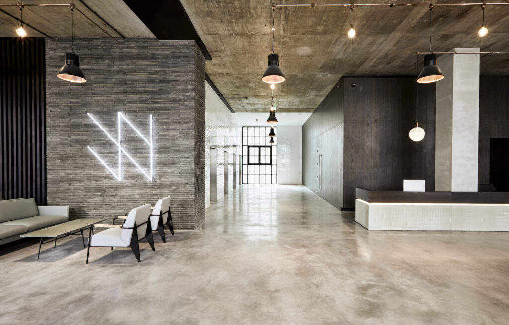An industrial-style interior of the Real PM Wenlock Works construction project in London. It features a modern slate-grey interior with pendant lights and a white neon sign.